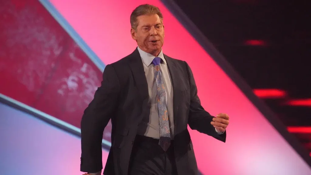 a5756be9-vince-mcmahon-investigation WWE Chairman Vince McMahon in black suit and patterned tie speaking at corporate event with pink and blue lighting