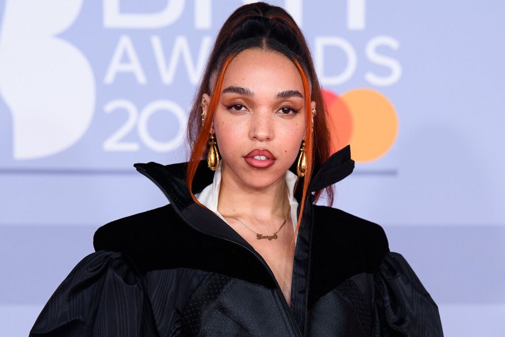 fka-twigs-1-fa00608488ed4609a75c0e8e8605c231 FKA Twigs at the BRIT Awards, wearing a high-collared black jacket with gold earrings and subtle makeup. Her long hair is styled in a high ponytail with orange streaks.
