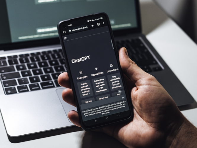 image-70_1745928790 Person holding a smartphone with the ChatGPT website open, positioned above a laptop keyboard. The screen displays ChatGPT’s interface with options like "Examples," "Capabilities," and "Limitations.