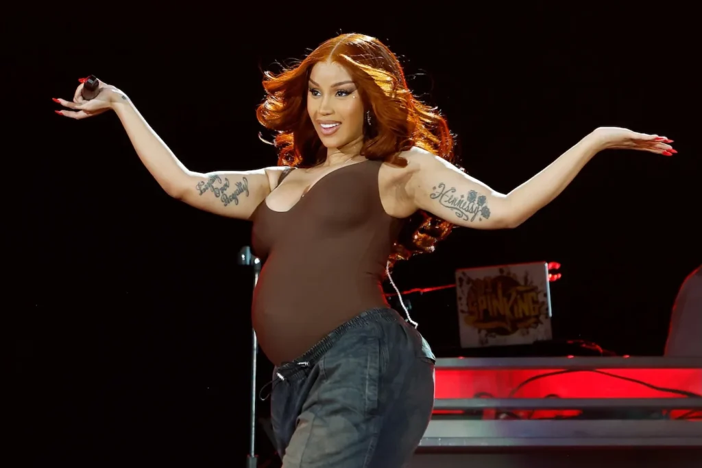 Rapper Cardi B on stage with arms outstretched, performing while visibly pregnant and showing a baby bump under a fitted brown top, stage lights glowing behind her.
